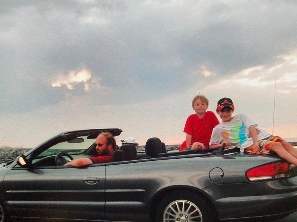 A man at the wheel of a convertible with two boys sitting up on the back seat, under a cloudy evening sky