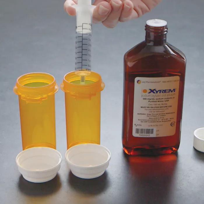 A bottle of Xyrem (sodium oxybate oral solution, 500 mg/mL) beside two pharmacy vials and a dosing syringe marked in grams.